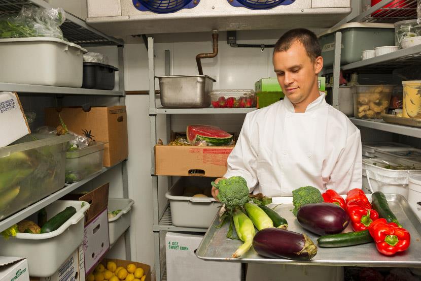 man in white chef coat examining tray of vegatables in a commercial walk in fridge