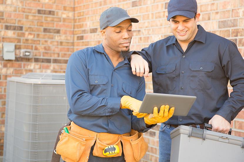 two men in blue standing in front of hvac units and looking at a tablet